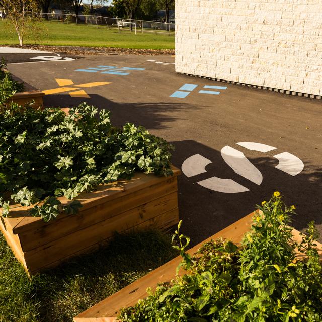 Lord Nelson School. Just outside the gym there are several wooden planters filled with lush plants. Colourful stencils of giant numbers and letters can be seen on the pavement. 